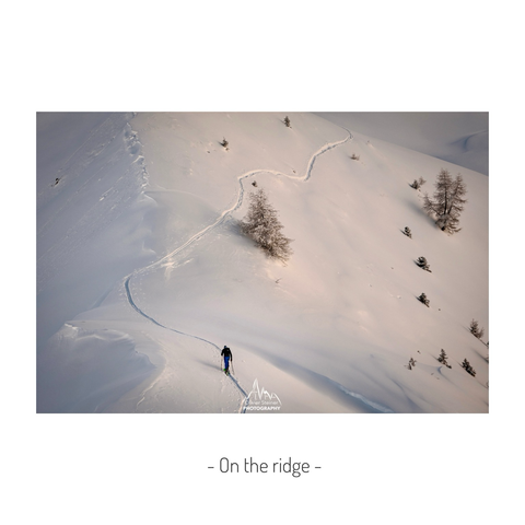A skier moves along a narrow snow-covered ridge lined with a few scattered trees. Soft light highlights the winding track in the snow, while the surrounding mountains and sky create a calm winter atmosphere. The scene emphasizes solitude and the scale of the landscape.