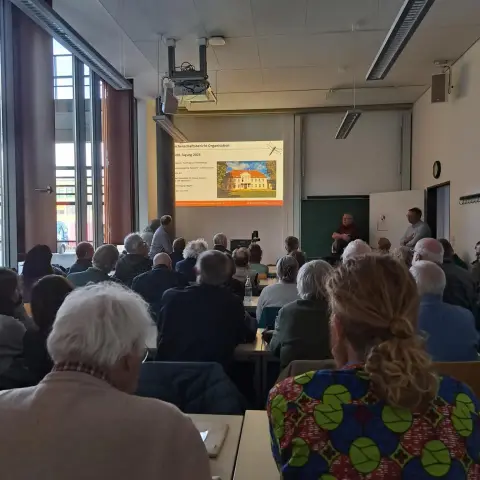 Ein langgestreckter Seminarraum an einer Universität. Viele Menschen sitzen an Tischen, mit dem Rücken zum Fotografen, und schauen auf eine Leinwand, auf der eine Tagungsfolie mit Text und dem Bild eines Herrenhauses zu sehen ist.