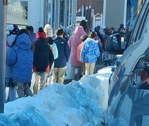 A line of children walking down a sidewalk next to a 2 foot high snowbank, some talking and walking together