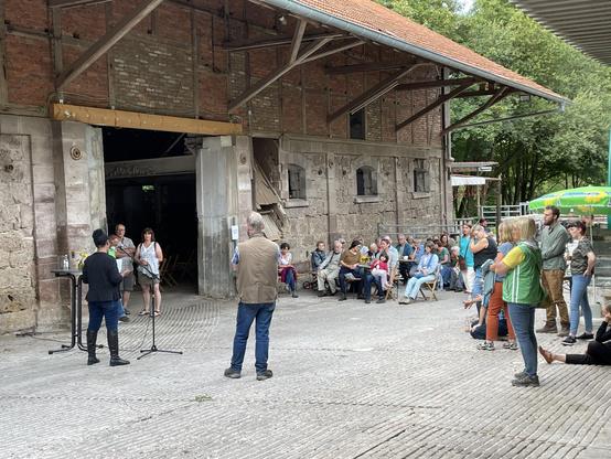 Das Foto zeigt eine öffentliche Veranstaltung im Freien auf einem gepflasterten Hof vor einem alten, teilweise offenen Gebäude aus Stein und Ziegeln, vermutlich eine ehemalige Scheune oder ein Wirtschaftsgebäude. Das Gebäude hat hohe Mauern, sichtbare Holzbalken unter dem Dach und große Öffnungen, durch die man ins Innere sehen kann.

Im Vordergrund stehen mehrere Menschen, die einer Ansprache zuhören. Links stehen zwei Personen an Mikrofonständern, es ist Awet. Sie hält Notizen in der Hand. Davor, mit dem Rücken zur Kamera, steht eine weitere Person, die dem Geschehen zuhört.

Rechts und mittig im Bild sitzt und steht das Publikum: Erwachsene unterschiedlichen Alters sowie einige Kinder. Einige sitzen auf einfachen Stühlen oder Bänken, andere stehen locker beisammen. Die Menschen wirken aufmerksam und entspannt, teils mit verschränkten Armen, teils im Gespräch oder nach vorne blickend.

Im Hintergrund sind Bäume und Grünflächen zu sehen, was auf eine ländliche oder parkähnliche Umgebung hindeutet. Rechts außen erkennt man einen Sonnenschirm, der auf eine Bewirtung oder ein Fest hindeutet. Insgesamt vermittelt das Bild den Eindruck einer ruhigen, gemeinschaftlichen Kultur- oder Informationsveranstaltung in historischem Ambiente.