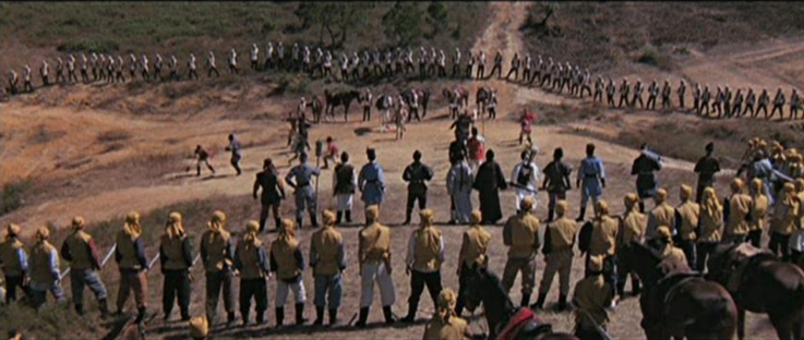 an image from the film, it is of many warriors standing in a large circle at the top of a hill, just small shrubs and grasses, some horses in the foreground with men holding their reigns 
