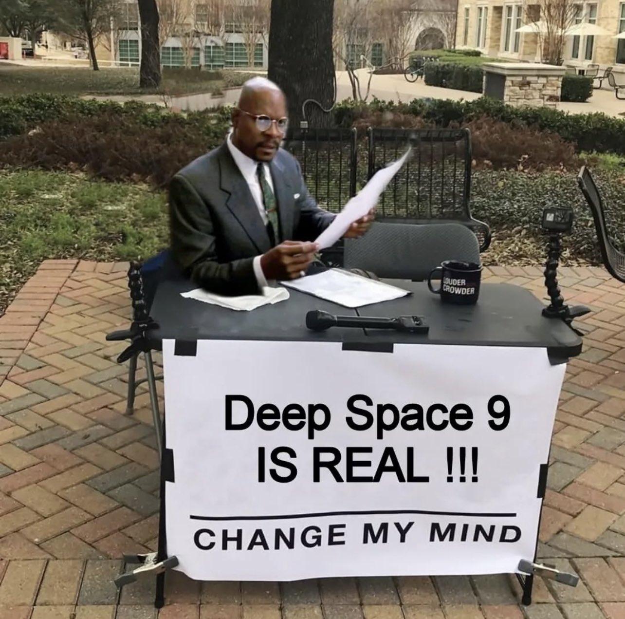 Benjamin Sisko sits at a table in a park dressed up as Benny from the DS9 episode Far Beyond The Stars. The sign taped to the front of the table reads: Deep Space 9 IS REAL!!! CHANGE MY MIND