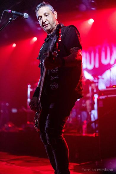 A musician plays an electric bass guitar on stage under red lighting, wearing a black vest with patches and a shirt. The atmosphere suggests a live performance.