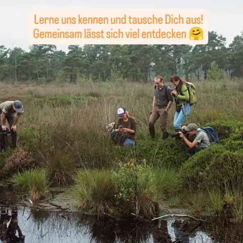 Eine Gruppe Menschen, die meisten mit einer Fotokamera oder Smartphone in der Hand, stehen am Rand eines Moorgewässers. Im Hintergrund Kiefern unter bedecktem Himmel. Text: Lerne uns kennen und tausche dich aus! Gemeinsam lässt sich viel entdecken (Umarmungs-Emoji)