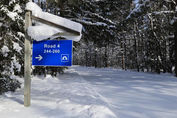 This is a landscape format photo taken in the winter of a snow covered campground road that has been groomed for cross country skiing. The ski trail passes through a forest and is quite flat in this section. A campground road sign that is blue and white in colour hangs from a wooden support which is also topped with snow. Many of the tree branches are covered in snow, which provides a real winter vibe for this photo.