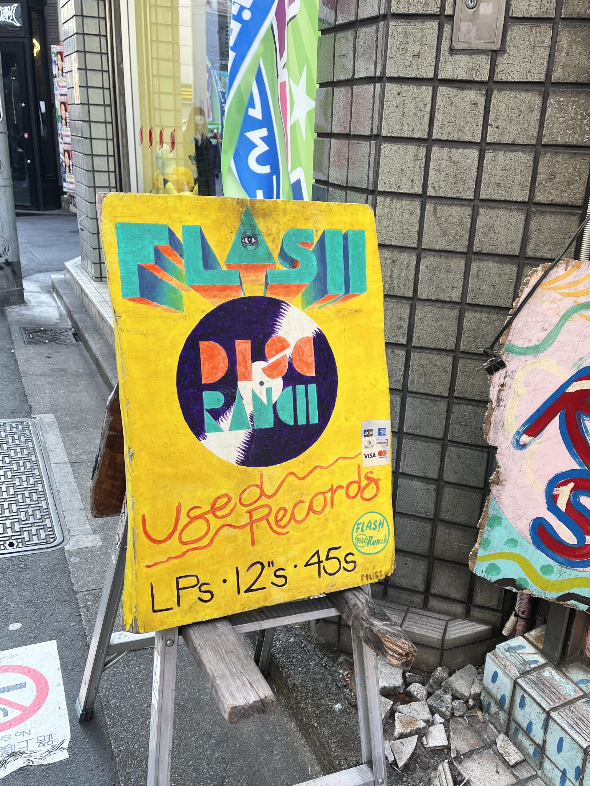 A bright yellow handmade sign for Flash Disk Ranch at the bottom of the stairs on the street in Shimokita.