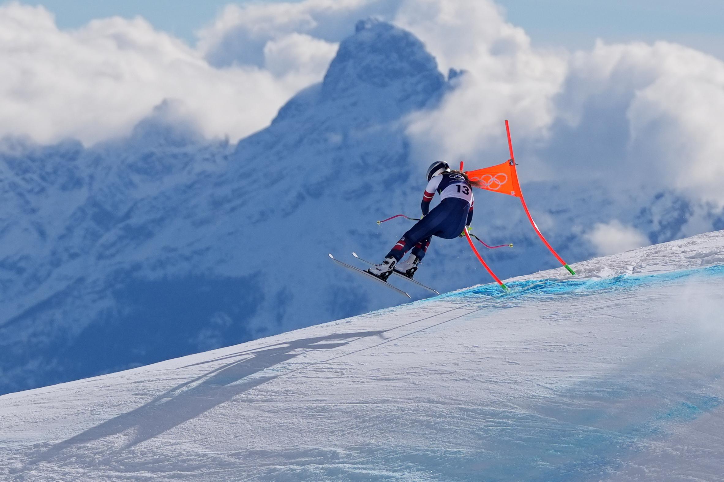 Photograph of Lindsey Vonn skiing in the downhill, partly in mid air with her arm around the pole of one of the gates on the course. Clouds and majestic mountains are seen in the background.