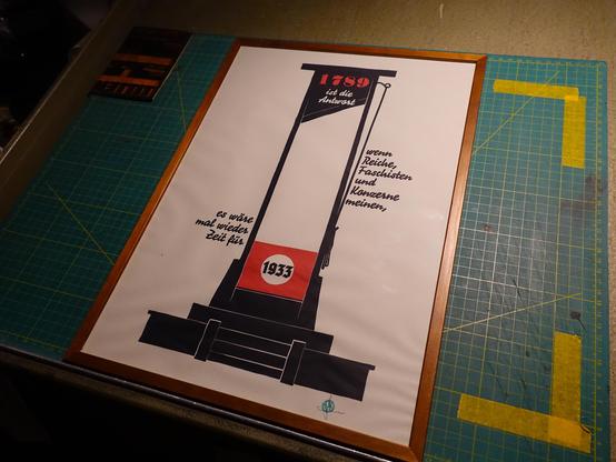 Foto eines gerahmten, hochformatigen Druckes, auf dem eine schwarze Guillotine zu sehen ist. Oben an ihrem Kopfende steht 1789 in rot, darunter in weiß ist die Antwort, links und rechts als Textfahnen wird der Satz fortgeführt: Wenn Reiche, Faschisten und Konzerne meinen, es wäre mal wieder Zeit für. Und im Kopfloch steht 1933 in schwarzer Fraktur. Das Kopfbrett der Köpfvorrichtung ist rot, so dass es wie eine stilisierte NSDAP Fahne aussieht.