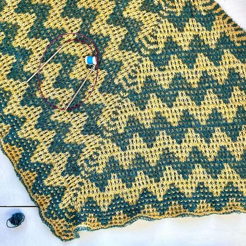 A flat lay view of a shawl with chevron mosaic patterning in teal and yellow on a white plank background. The shawl is finished, but not yet blocked.