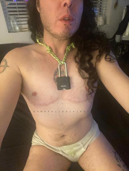 a light skinned ftm with long dark curly hair and short chin stubble wearing a lime green rope closed with a large padlock around his neck as a symbol of his submission and tighty whitey briefs does a selfie on his bed.