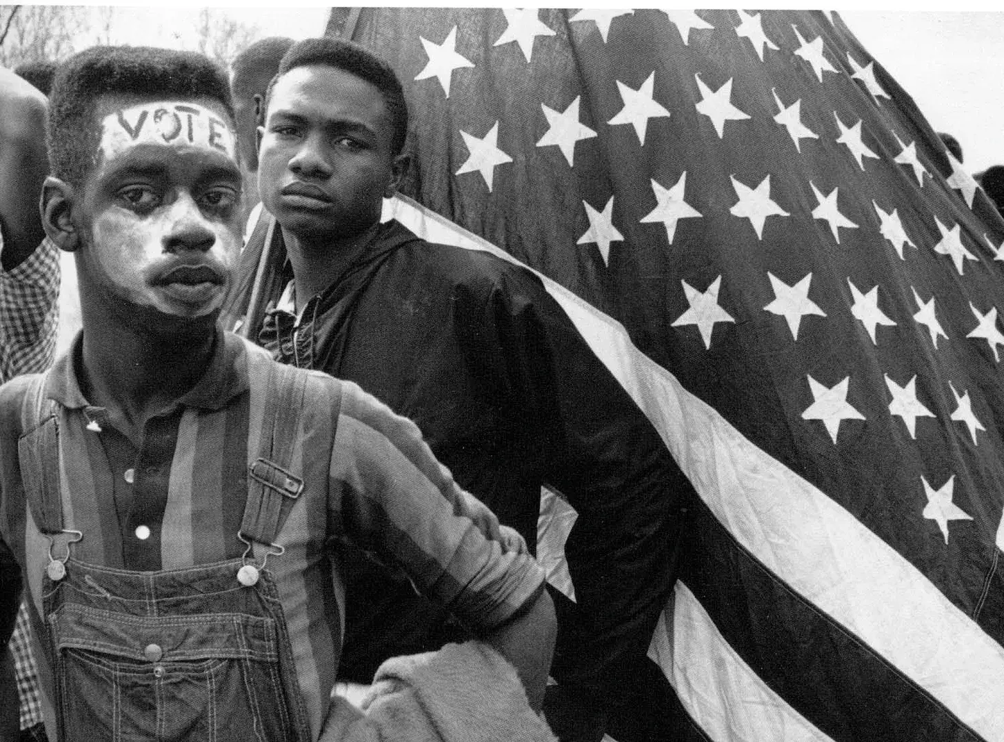 A close-up, black-and-white photograph by Bruce Davidson depicts Bobby Simmons, a young African American man, during the 1965 Selma to Montgomery march. He gazes directly at the camera with a somber, determined expression, his face marked by the word "VOTE" written in dark letters across his forehead. His cheeks and chin are covered in a thick layer of white zinc oxide, used by marchers for sun protection. Wearing striped overalls, he stands in the foreground as another young marcher holds a large, draped American flag behind him, creating a powerful contrast between the symbols of national identity and the personal struggle for civil rights.