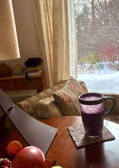 Adrian's breakfast smoothie sitting in a blender mug on a table in his home, together with some fruit and a table lamp. Beyond a brightly upholstered chair, a snowy Vermont winter scene is visible outside.