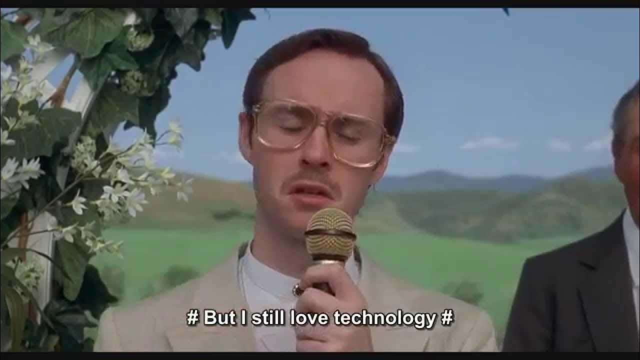 Still from Napoleon Dynamite: Guy in a suit holding mic. He's singing. "But i still love technology"