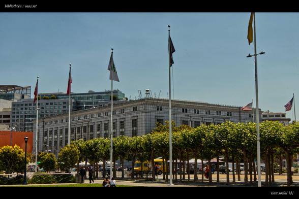 A color photo of the Central Branch of the San Francisco Public Library system with flags in the foreground and commercial building in the background.