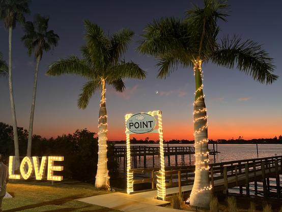 Photo by Fenichel:
Palm trees and a dock are in the foreground, water and vivid sunset in the rear.
To the left is a big lit sign saying "LOVE" and in the center, entrance to the dock, is a sign that says, "The POINT". 