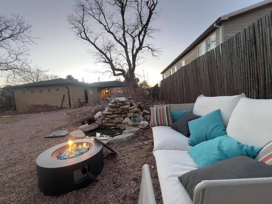 Twilight, clear sky above a blonde brick backend of a house in the distance; silhouette of a leafless tree reaches into the sky. Sandstone slabs form a fountain, water falling over them into a small round pool. A circular metal fire pit contains some flames, with a white cushioned couch next to it all. 