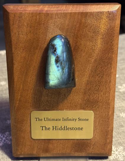 Reddish wooden board, polished and oiled with an embedded, blueish.green shimmering Labradorite and a brass plate underneath, reading "The Ultimate Infinity Stone - The Hiddlestone".