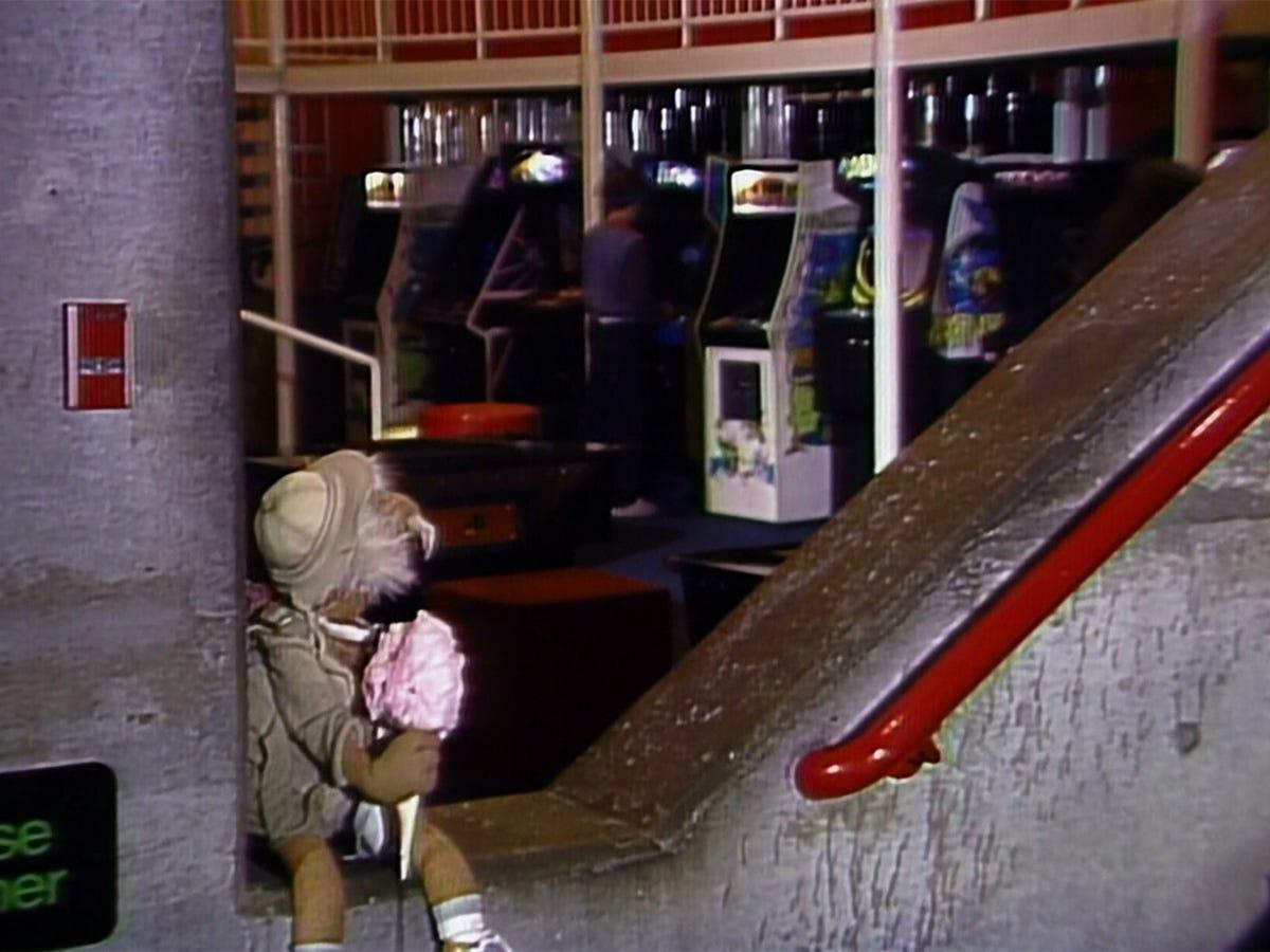 Fraggle Rock's Traveling Matt sitting on a great concrete half wall holding pink cotton candy while looking lovingly at an arcade