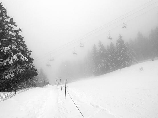 blanc and white picture of an alpine ski piste, bordered with snowy conifer that disappear into the fog. A chairlift crosses the picture above the piste.