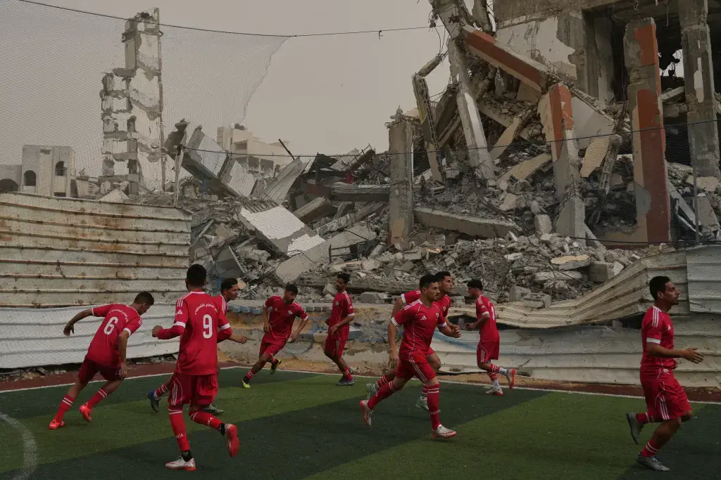 Palestinian players from Al-Ahly club warm up before a football match at a newly constructed field surrounded by destroyed buildings.