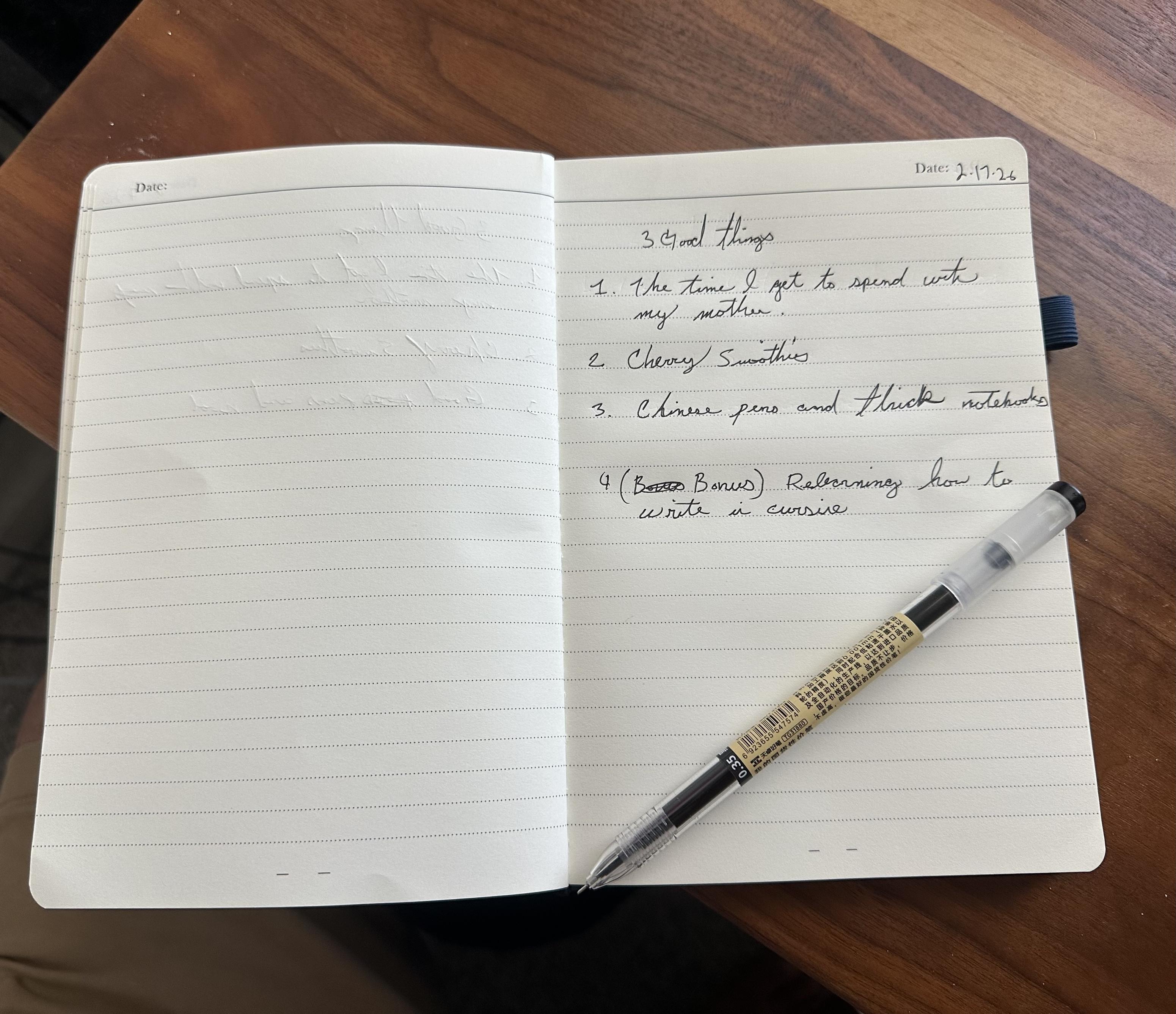 My notebook, open, with a pen laying on the page. Date 2/16/26 It's reads, in cursive, illegibly: 3 Good things 1. The time I get to spend with my mother 2. Cherry smoothies 3. Chinese pens and thick notebooks 4. (Bonus) Relearning how to write in cursive