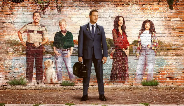 Photo of lead actor with medical bag, in front of brick wall with painted photos of four other actors and a small white dog