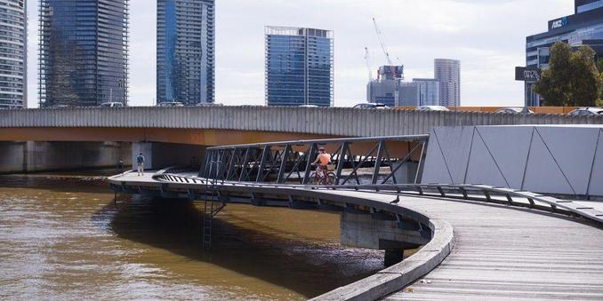 Photo of a curved, wood-and-metal, pedestrian and cyclist bridge that runs along the length of an urban river. The walkway and cycle path along the river have been interrupted by a road bridge that spans the river. This pedestrian and cyclist bridge connects these interrupted paths via a curved section that goes below the road bridge.