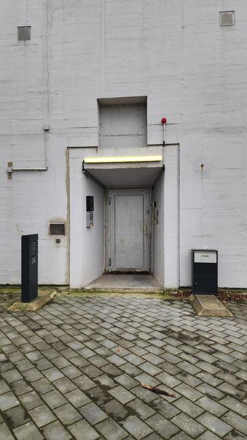 An iron door, an entry into an office and flat, as well as a former air raid shelter. There is a black rectangular, small obelisk to the left and another, shorter one to the right of the entry. The one to the left has the housenumber 34 on it. The other seems to be a letterbox.