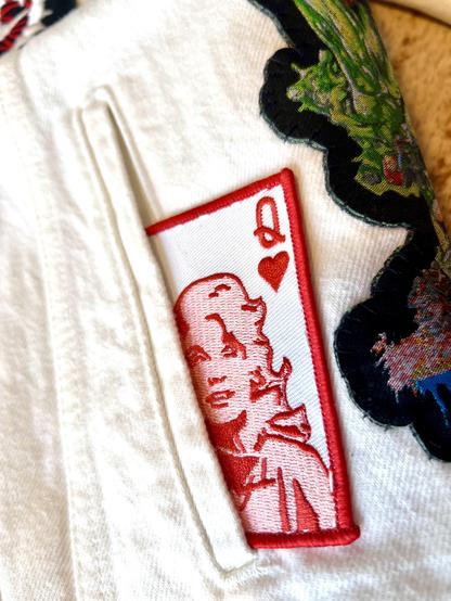 A Dolly Parton queen of hearts patch sewn half in and half outside of a white denim battle jacket pocket. 