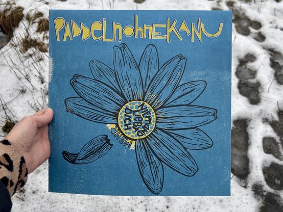 Ein Albumcover mit eines Linolschnitts eines Gänseblümchens auf blauem Grund. Ein paar Blütenblätter fehlen, eines löst sich gerade vom Blütenstandsboden. Die inneren, gelben Röhrenblüten sind angedeutet in gelb auf den blauen Untergrund gedruckt. Der Schriftzug paddelnohnekanu ist gelb mit weißer Umrandung prominent am oberen Rand platziert. Daraus ergibt sich eine Farbpalette von blau, gelb, weiß und schwarz für das Cover. Eine Hand hält das Album vor einem Hintergrund aus Schnee mit Fußspuren.