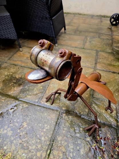 Ein aus diversen Stahlresten zusammengeschweißter Drache steht im Regen auf meiner Terrasse. Die Oberseite seines Schnabels ist ein Stück eines großen Esslöffels. Der Rest des Wesens ist überwiegend mit Rost überzogen, da er aus Baustahl, verschiedenen Rohren, Ketten, Schellen und Schraubenschlüsseln besteht.
