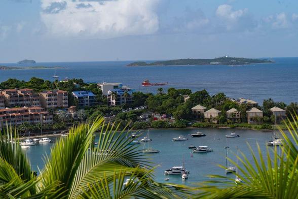 View of Cruz Bay on St. John in the U.S. Virgin Islands, with sailboats anchored in turquoise water below, palm fronds framing the foreground, and small green islands layered across the blue horizon.