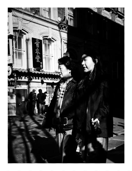 Black and white street photograph of two Chinese women walking in dappled sunlight on George Street in Manchester's Chinatown, with traditional architecture and other pedestrians visible in the background.