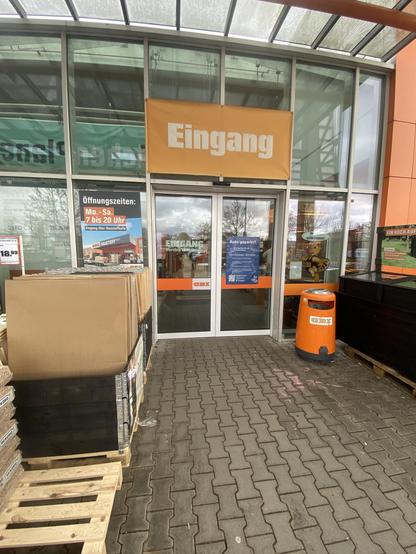 Image of a store entrance with a large "Eingang" sign. There is a poster displaying opening hours and a trash bin nearby. The area is paved with bricks and has some wooden pallets with products stacked beside the entrance.