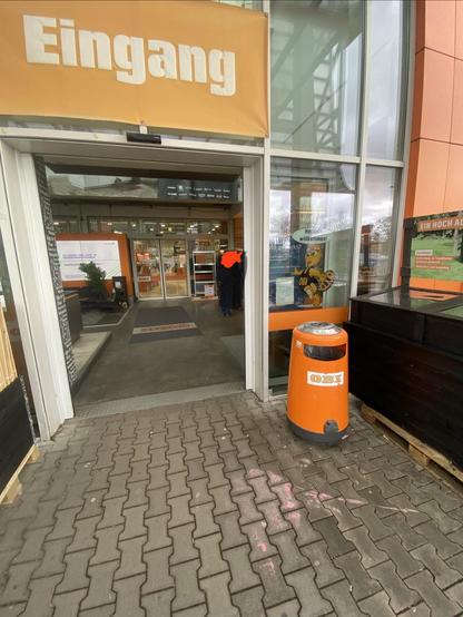Entrance of a store with a prominent "Eingang" sign. An orange waste bin is visible on the right, and there are people inside the store. The floor features patterned pavement, and a welcoming poster is partially visible through the entrance.