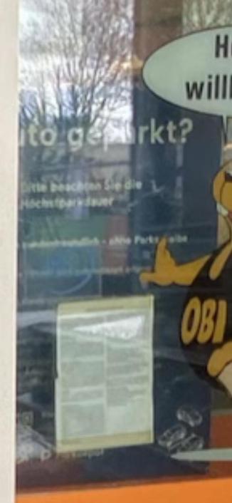 The image shows a window display featuring a cartoon character in a vivid outfit with the text “Wie wird geparkt?” and other German text. There is also a document visible behind the glass with additional text. The background includes trees and what appears to