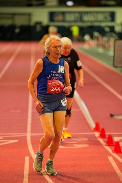 In the front a woman with short white hair wearing a blue tank top and grey shorts with bib number 244 and a white rectangle with the number 10 n her shoulder is in focus,  the runners behind her including a woman in a black shirt are blurry,  the background is a red track with a blurred sig that says NAVY / MARINES next to an American flag in the far background