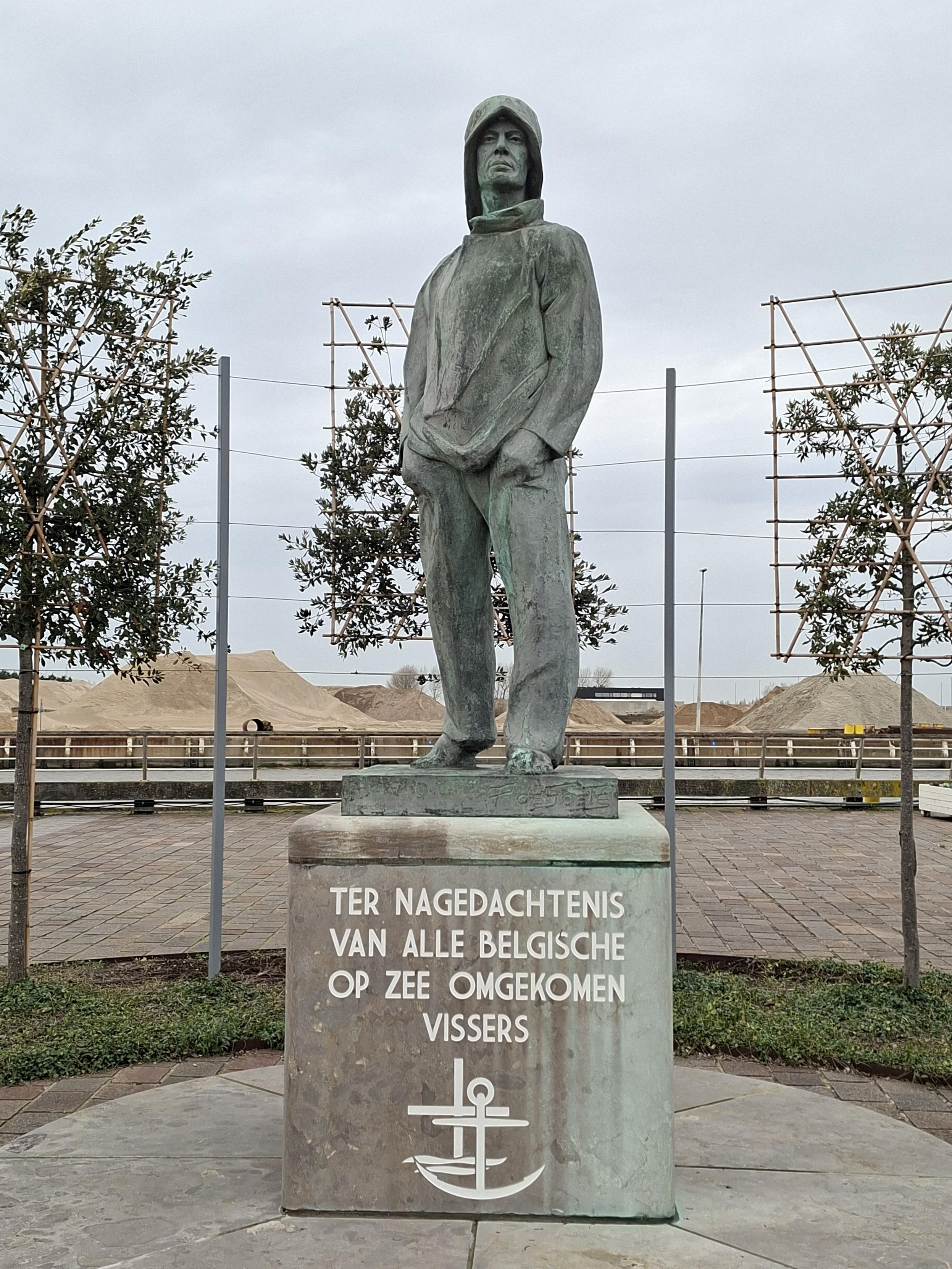 A statue of a fishermen in memoriam of  those who died at sea. Behind it are small trees and a bit further are sand piles ready to be shipped.
