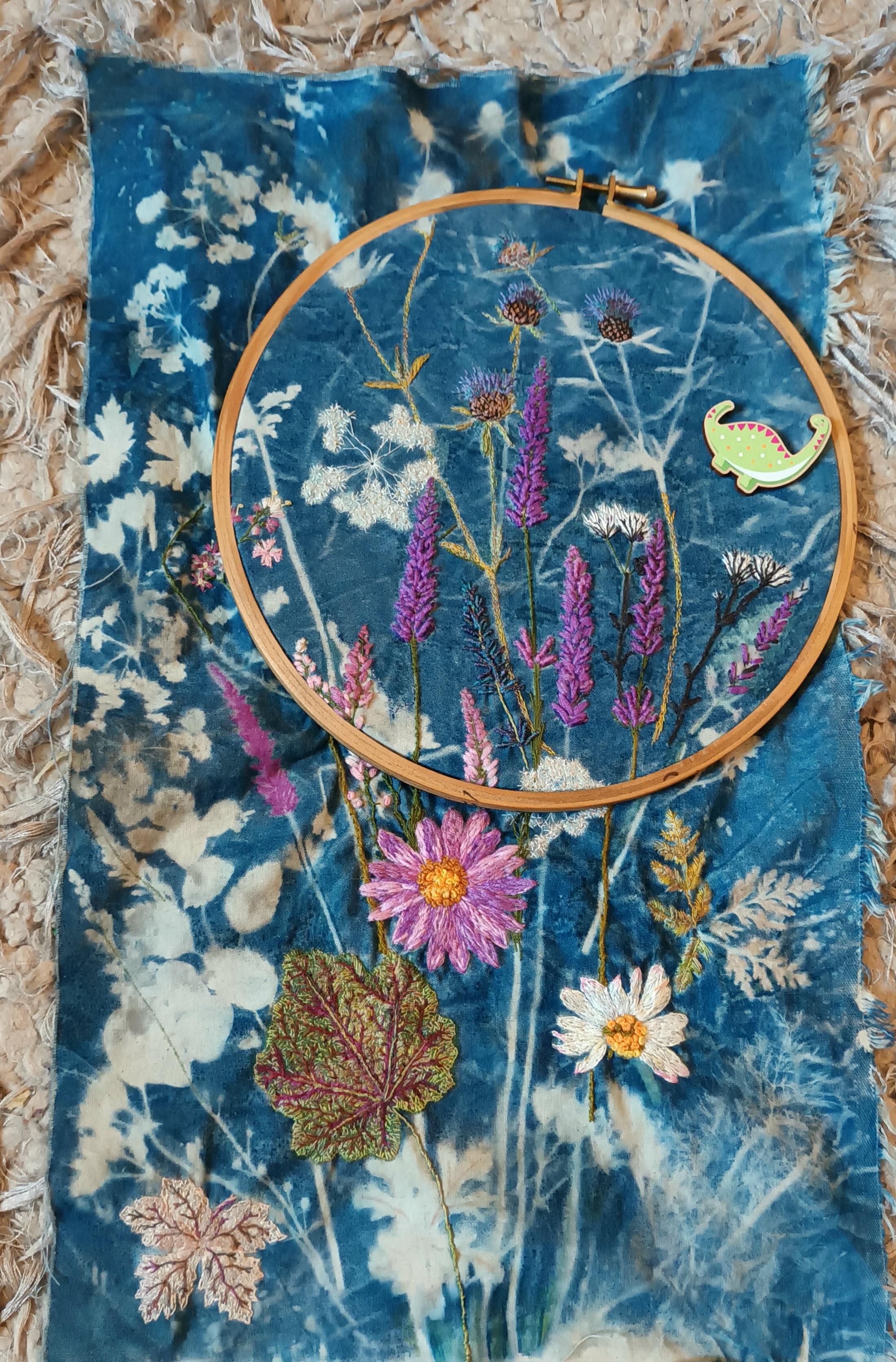 Photograph of a cyanotype print on cotton with hand embroidery over the top, in places. The print was made using flowers that were in bloom in September and include lavender, heuchera, ox eye daisy, toadflax and knapweed.