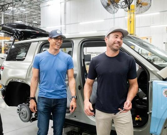 Rivian CEO RJ Scaringe & CrunchLabs founder Mark Rober stand in front of an R1S EV half assembled on the Rivian assembly line. 