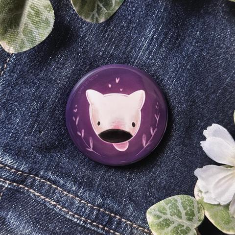 Picture of a round button badge pinned on a jean jacket, framed with decorative plants. The badge features a digital illustration of a cute white bull terrier head floating on a purple background. 