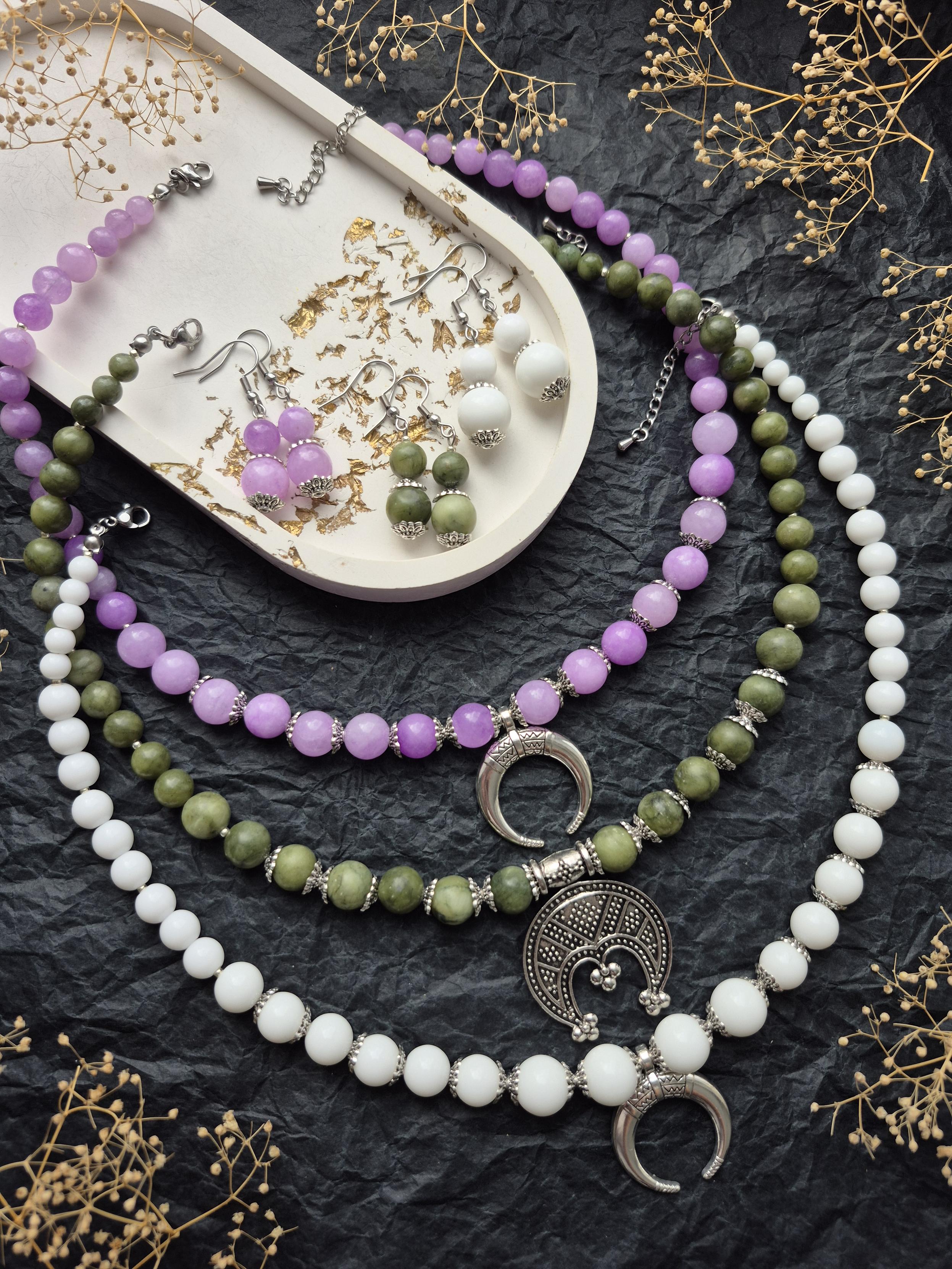 A collection of handmade iewelrv on a black textured background. Three necklaces made of natural stones (delicate lilac, olive green, and milky white) are decorated with silver moon pendants in ethnic style. Nearby, on a decorative light tray, lie three pairs of matching earrings The composition is complemented by sprigs of dried flowers.
