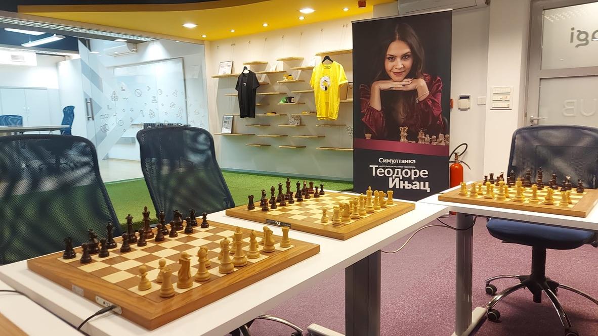 Chess boards set up on tables with office chairs at the Kagi Hub venue, ready for the Rising Stars of Chess 2026 simultaneous exhibition. A promotional banner in Serbian featuring a portrait of chess champion Teodora Injac stands between the boards. Kagi-branded merchandise including black and yellow t-shirts is displayed on wall shelves.