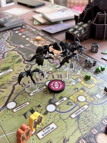 A tabletop game scene featuring a detailed game board with various locations. Six black Nazgûl miniatures are positioned on clear stands, near a the eye of Sauron token. Various cards and game pieces are scattered around, indicating an active gameplay setting.