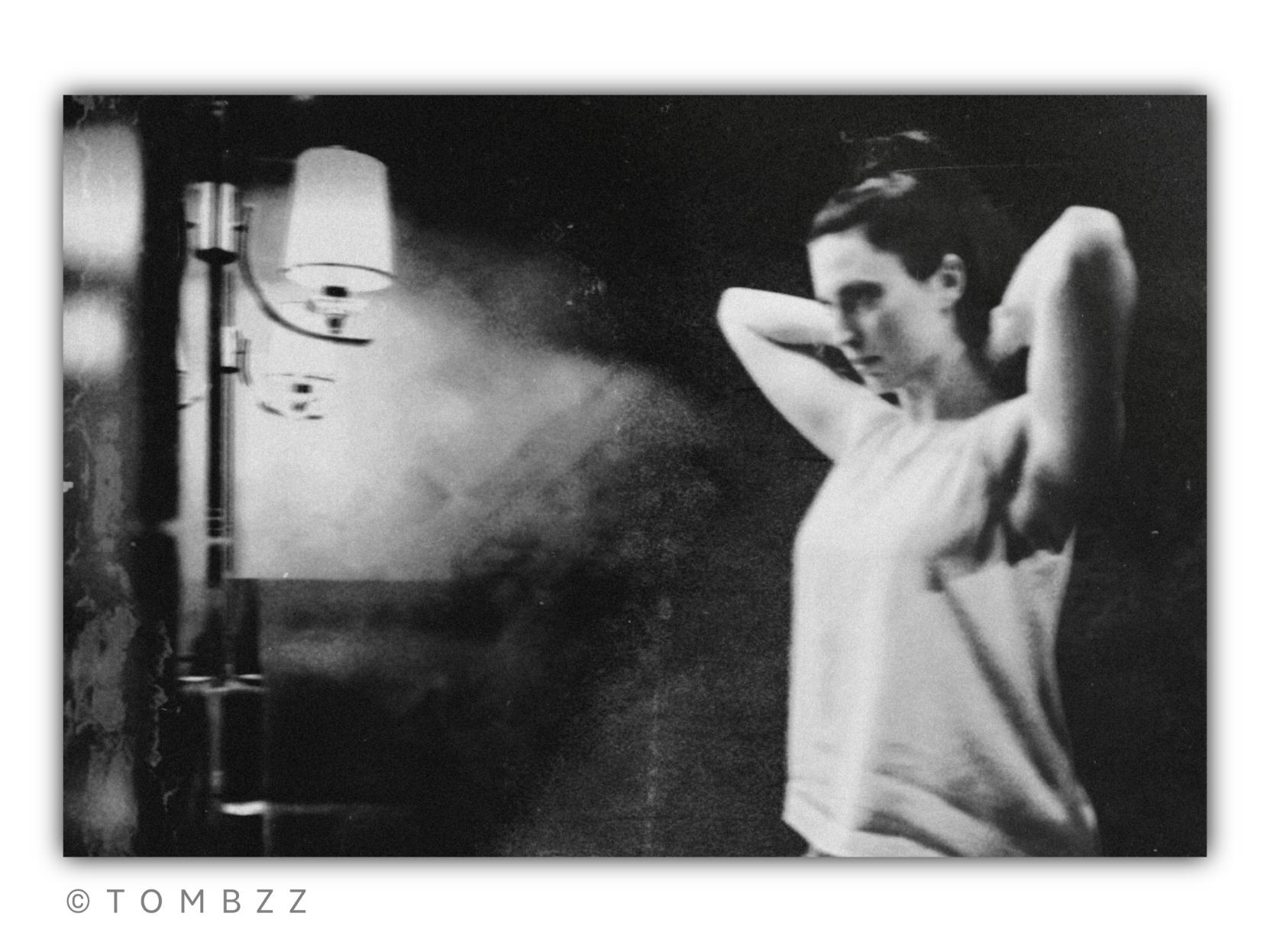 A black and white analog photograph that, through its grain and play of shadows, directly references the aesthetic of film noir and the stifling atmosphere of the Cinema of Moral Anxiety. The frame is dense, intimate, and full of understatements.
​On the right side, a young woman with dark hair is captured in profile, frozen in the gesture of adjusting her hair. Her arms are raised, creating a triangular frame around her face. She does not look at her hands but fixes her gaze on a mirror invisible to us. This look is not one of vanity; it is piercing, focused, and heavy with suppressed emotions, as if she is searching her reflection for an answer to a question she is afraid to ask aloud.
​The left side of the frame is dominated by the soft, directional light of a wall lamp with a milky shade. It pulls the texture of her light-colored blouse and the delicate silhouette from the darkness, while the rest of the room is submerged in deep, grainy blacks. The composition suggests a moment of suspension – a brief heartbeat before an irreversible decision or stepping out into the night. The scene, though seemingly mundane, emanates a premonition of something unexpected about to happen just beyond the edge of the photograph.
