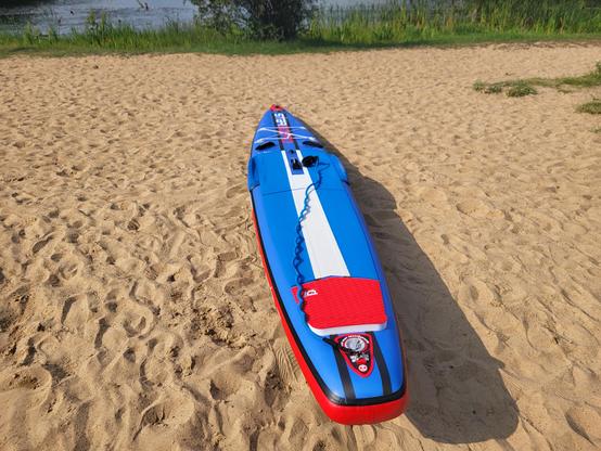 Ein blau rot weißes SUP was auf Sand an einem See liegt. Im Hintergrund sieht man noch etwas Wasser. Am Ufer befindet sich Gras.