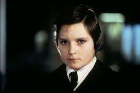 Promotional image of the character, Damien, from the movie Omen II. He looks to be about 12-15 years old. He has a stern expression on his face. Dark combed back hair. Unsmiling. Wearing a dark suit, white shirt and tie. The resemblance between the character and Baron Trump is striking. 