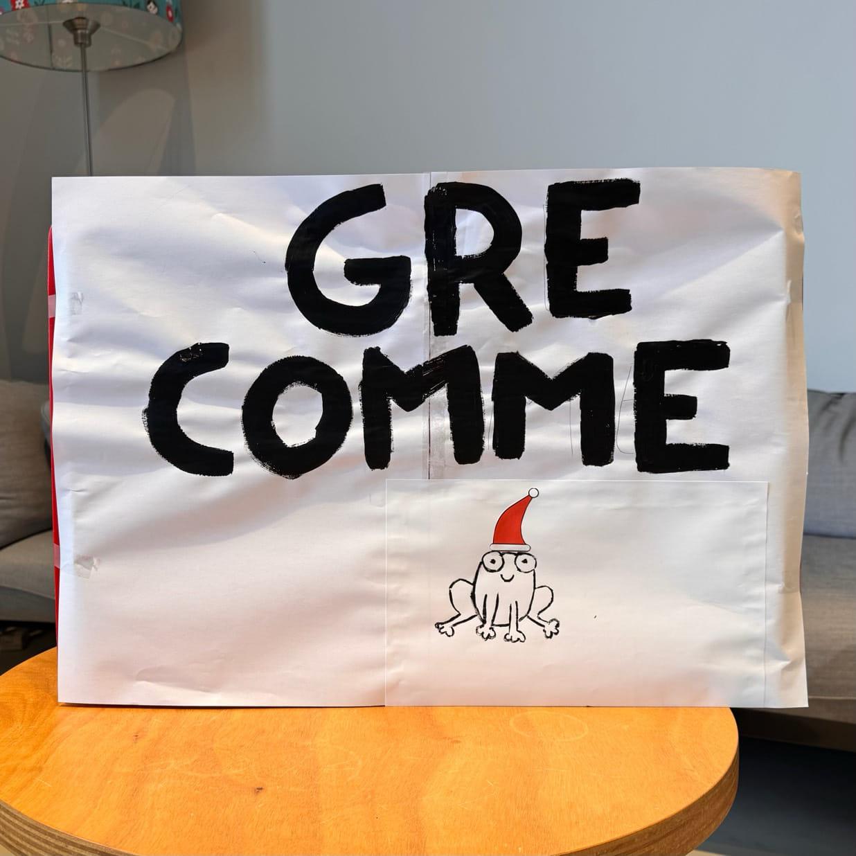 Une affiche, peinte à la main, disposée à la verticale sur une table en bois. Il est écrit "GRE COMME" et dessous, est peinte une grenouille aux formes naïves et à l