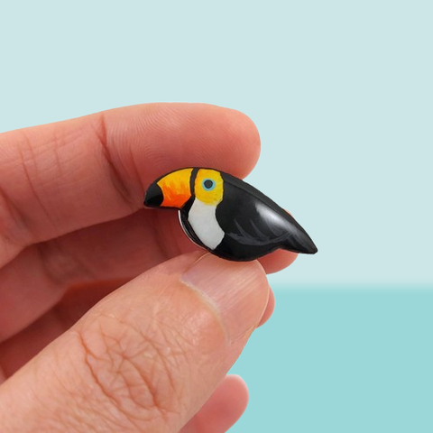 Pin's éco-responsable en forme de petit toucan toco.
La perle est réalisée en CD recyclé, découpée et peinte à la main, puis recouverte de résine.
Il est tenu entre l'index et le pouce d'une main, devant un fond bleu pastel.