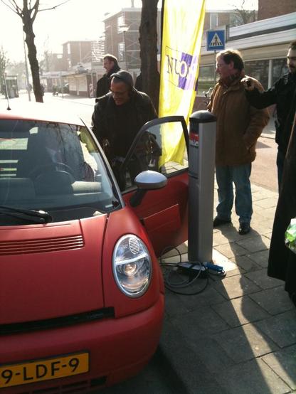 Mensen rond een rode Think City elektrische auto, naast een oplaadpunt. Erbij een gele vlag met paars Nuon-logo.